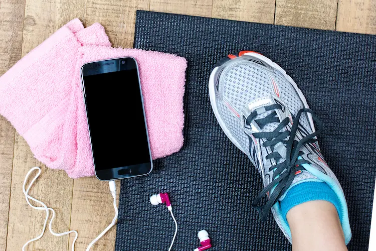 person in tennis shoe with phone earbuds towel and workout mat
