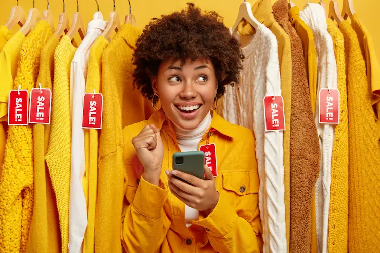 woman with phone in clothing wrack with sale tags stop buying clothes