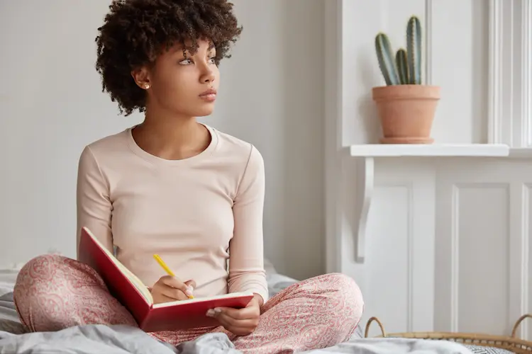 woman sitting on bed writing in journal looking out thinking